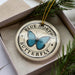 A circular ornament with a blue butterfly design and the text "BLUE MORPHO BUTTERFLY" printed on it, placed inside a box with pine branches.