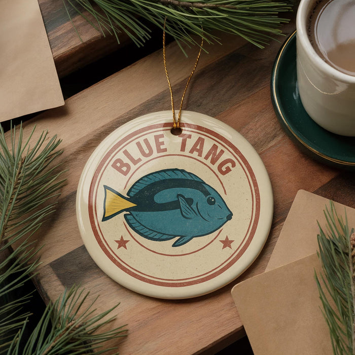 A circular ornament with a blue fish design and the text "BLUE TANG" is displayed on a wooden surface, surrounded by pine branches and a cup of coffee.