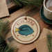A circular ornament with a blue fish design and the text "BLUE TANG" is displayed on a wooden surface, surrounded by pine branches and a cup of coffee.