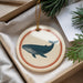 A round ornament featuring a blue whale is displayed in a box, surrounded by pine branches and a white card.