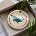 A decorative ornament in a box, featuring a whale silhouette on a white background with a red border.
