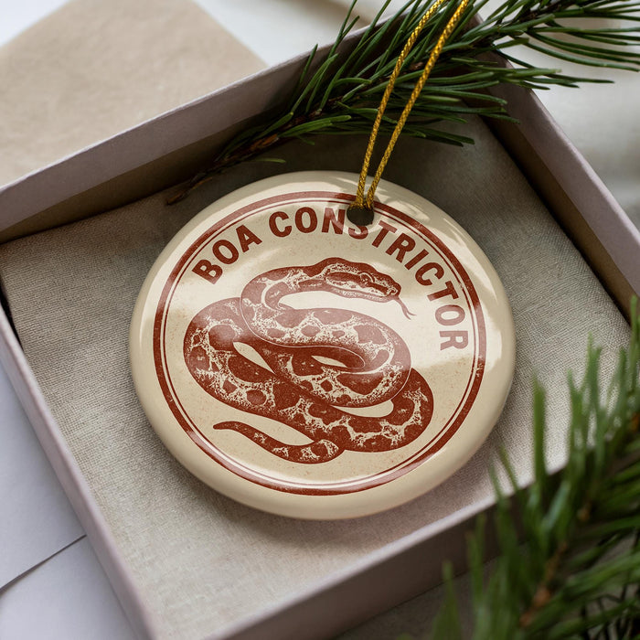 A round ornament with the text "BOA CONSTRICTOR" and a snake design, placed inside a box with pine branches.