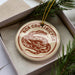 A round ornament with the text "BOA CONSTRICTOR" and a snake design, placed inside a box with pine branches.