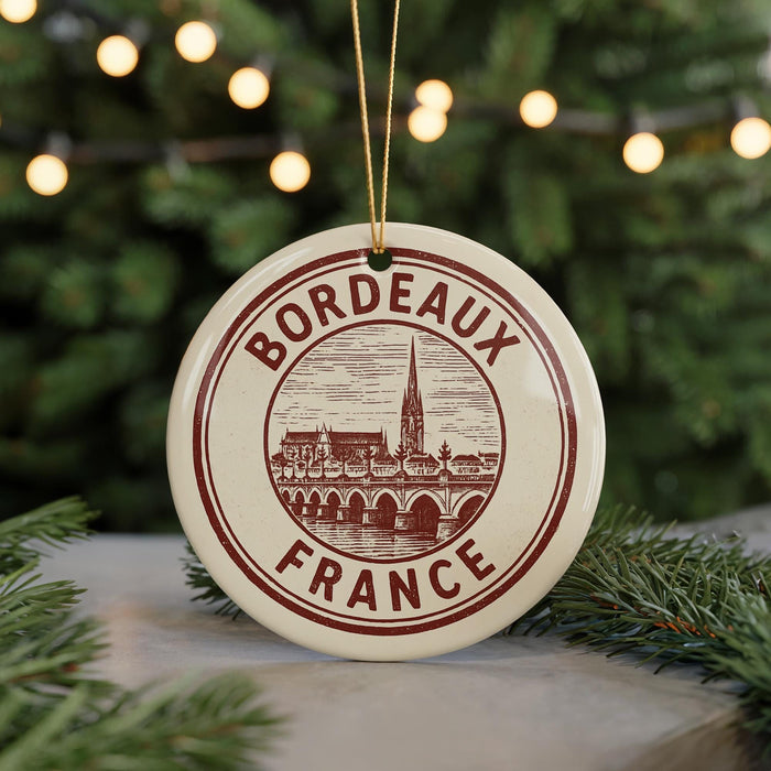 A round ornament with the text "BORDEAUX FRANCE" and an illustration of a bridge is displayed in front of a Christmas tree with twinkling lights.