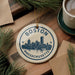 A circular ornament with the text "BOSTON MASSACHUSETTS" and a graphic of the Boston skyline, hanging on a wooden surface.