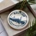 A circular ornament with the text "BOSTON MASSACHUSETTS" and a cityscape illustration, hanging from a string.