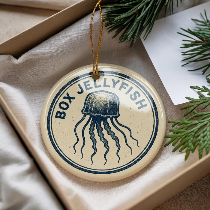 A round ornament with a blue jellyfish design and the text "BOX JELLYFISH" is displayed in a box, surrounded by pine branches.