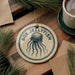 A round ornament with the text "BOX JELLYFISH" and an illustration of a jellyfish is displayed on a wooden surface, accompanied by pine branches and a cup of coffee.