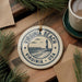 A decorative ornament with the text "VIRGINIA BEACH USA" and a lighthouse image, placed on a wooden surface next to a cup of coffee and some pine branches.
