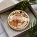 A circular ornament with a brown dog image and the text "Vizsla Dog" on it, placed in a box with pine branches.
