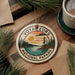 A decorative ornament with the text "VOYAGUEURS NATIONAL PAPARK" and a scenic landscape, featuring pine trees, a lake, and a sunset, is displayed on a wooden surface alongside a cup of coffee and some papers.
