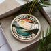 A circular ornament with the text "VOYAGE EURS NATIONAL PAPARK" and a landscape design, placed in a box with pine branches.