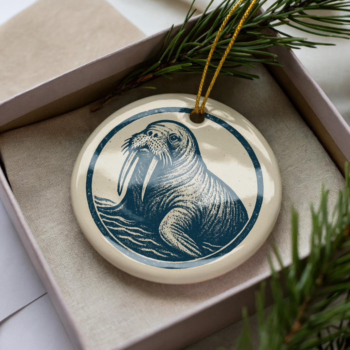 A ceramic ornament in the shape of a walrus, with a blue and white design, is displayed in a box with pine branches.