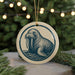 A circular ornament featuring a walrus with tusks, surrounded by a blue and white design, is displayed in front of a blurred Christmas tree with twinkling lights.