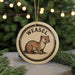A round ornament featuring a weasel, with the word "WEASEEL" written on it, is displayed in front of a Christmas tree with twinkling lights.