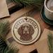 A circular ornament featuring a brown and white illustration of a Boykin Spaniel dog, with the text "BOYKIN SPANIEL" printed on it. The ornament is hanging from a string, and there is a cup of coffee and some pine branches in the background.