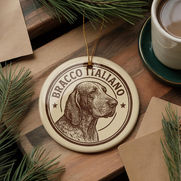 A circular ornament featuring a dog's head and the text "BRACCO ITALIANO" is displayed on a wooden surface, surrounded by pine branches and a cup of coffee.