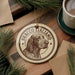 A circular ornament featuring a dog's head and the text "BRACCO ITALIANO" is displayed on a wooden surface, surrounded by pine branches and a cup of coffee.