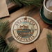 A circular ornament with the text "BRATSLAVA SLOVAKIA" and an illustration of a castle, placed on a wooden surface next to a cup of coffee and some pine branches.