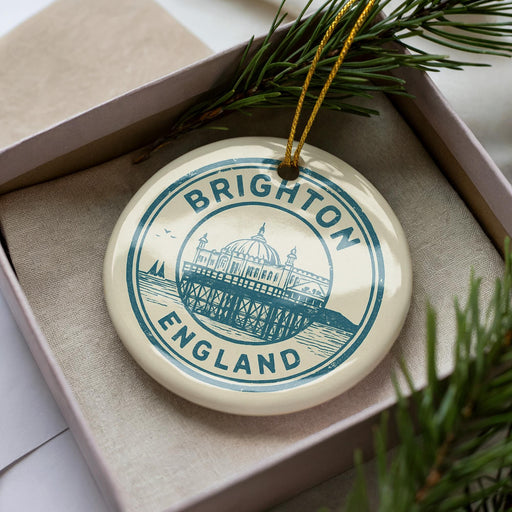 A circular ornament with the text "BRIGHTON ENGLAND" and an illustration of a building on a pier, placed inside a box with pine branches.