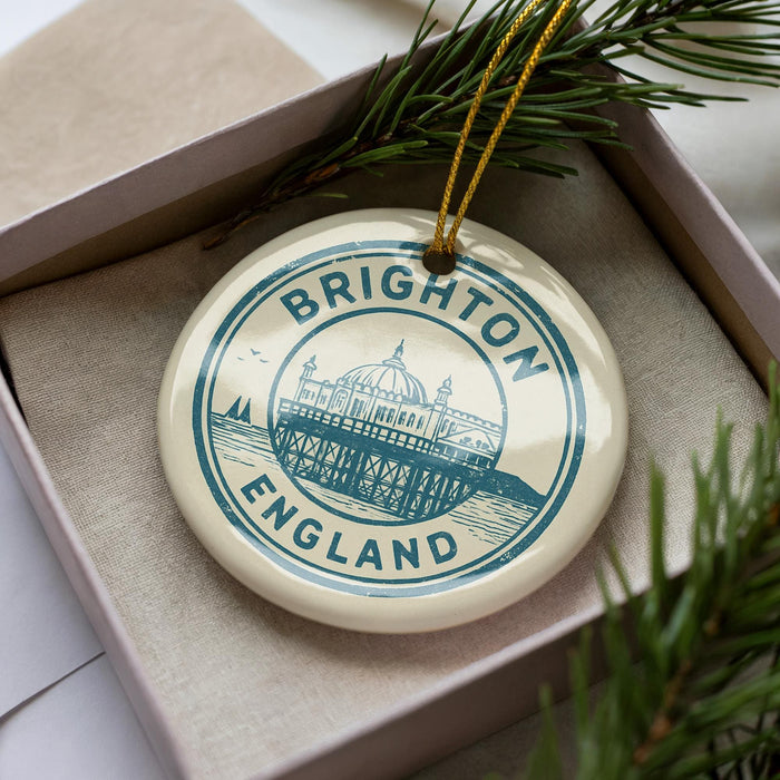 A circular ornament with the text "BRIGHTON ENGLAND" and an illustration of a building on a pier, placed inside a box with pine branches.