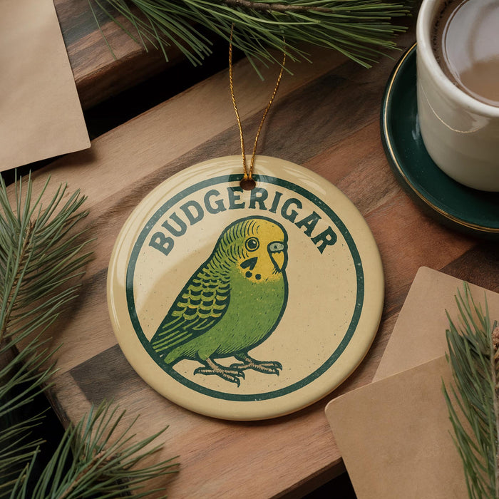A round ornament featuring a green parakeet, with the word "BUDGERIGAR" written on it, is displayed on a wooden surface alongside a cup of coffee and some pine branches.