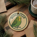 A round ornament featuring a green parakeet, with the word "BUDGERIGAR" written on it, is displayed on a wooden surface alongside a cup of coffee and some pine branches.