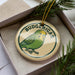 A circular ornament with a green parakeet design and the word "BUDGERIGAR" written on it, placed inside a box with pine branches.
