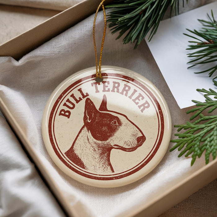 A bull terrier ornament is displayed in a box, surrounded by pine branches.