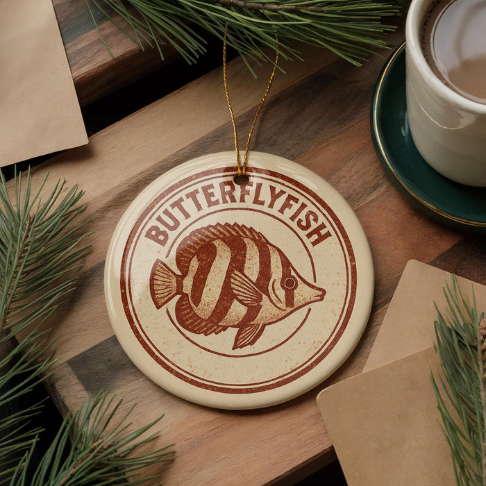 A round ornament with a butterflyfish design is displayed on a wooden surface, accompanied by a cup of coffee and some pine branches.