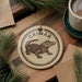 A circular ornament featuring a cartoon alligator is displayed on a wooden surface, surrounded by pine branches and a cup of coffee.