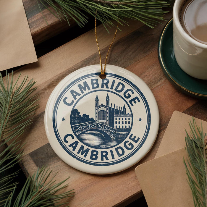 A ceramic ornament with the text "CAMBRIDGE" and an illustration of a bridge is displayed on a wooden surface, surrounded by pine branches and a cup of coffee.
