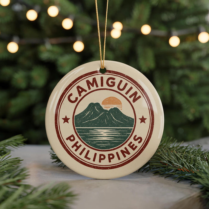A round ornament with the text "CAMIGUIN PHILIPPINES" and a graphic of a mountain and water, hanging from a string against a background of Christmas lights and greenery.