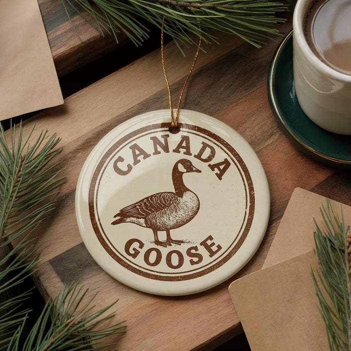 A circular ornament featuring a Canadian goose is displayed on a wooden surface, surrounded by pine branches and a cup of coffee.