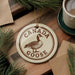 A circular ornament featuring a Canadian goose is displayed on a wooden surface, surrounded by pine branches and a cup of coffee.