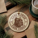 A ceramic ornament featuring a dog's head and the text "CANE CORSO" is displayed on a wooden surface, surrounded by pine branches and a cup of coffee.