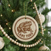 A Christmas tree decorated with ornaments, including a round ornament with a frog image and the text "CANE TOAD" printed on it.