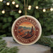A round ornament with a desert landscape design, featuring mountains and a sunset, is displayed in front of a Christmas tree with lights.