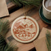 A decorative ornament featuring a coastal town scene with mountains, a boat, and the text "CAPRI ITALY" is displayed on a wooden surface, accompanied by pine branches and a cup of coffee.