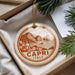 A decorative ornament with a circular design featuring a coastal town and mountains, along with the text "CAPRI ITALY". The ornament is hanging from a string and placed in a box with pine branches.