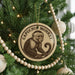 A round ornament with a monkey image is hanging on a Christmas tree, surrounded by other ornaments and lights.