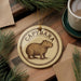 A round ornament with the word "CAPYBARA" printed on it, featuring an illustration of a capybara. The ornament is placed on a wooden surface, surrounded by pine branches and a cup of coffee.