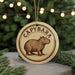A circular ornament featuring a brown capybara (or similar animal) on a beige background, with the word "CAPYBARA" written around the edge.