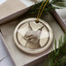 A ceramic ornament featuring a detailed illustration of a cat's face is displayed in a box, surrounded by pine branches.