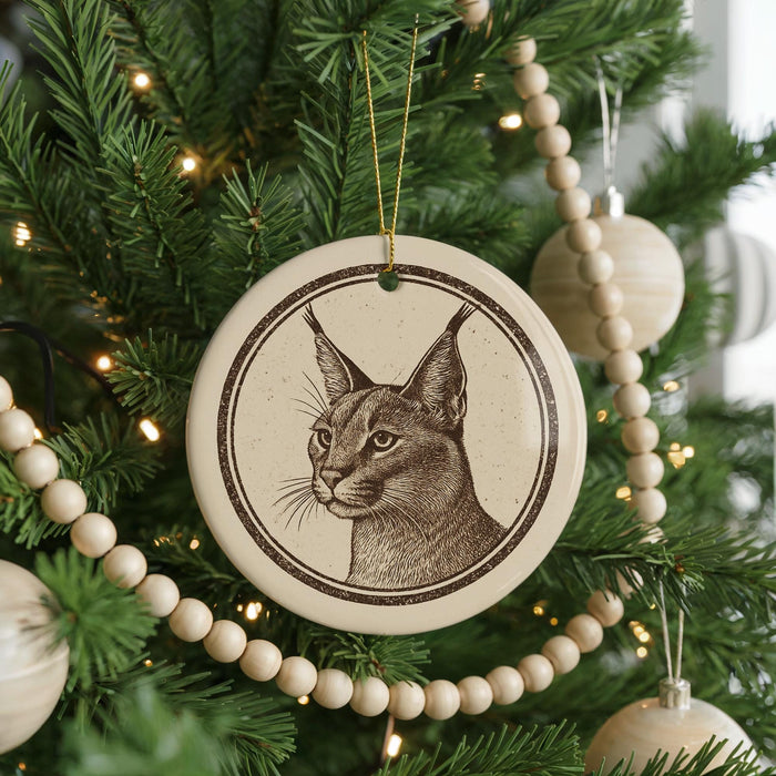 A ceramic ornament featuring a detailed illustration of a cat's face is hanging from a Christmas tree, surrounded by ornaments and lights.