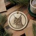 a ceramic ornament with a detailed illustration of a cat's face, placed on a wooden surface next to a cup of coffee and some pine branches.