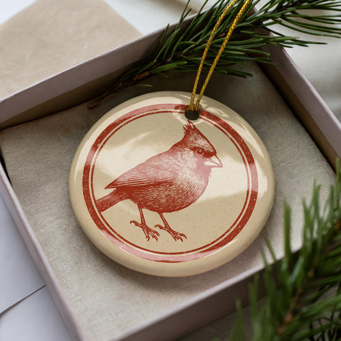 A red and white ceramic ornament featuring a detailed illustration of a cardinal bird is displayed in a box, surrounded by pine branches.