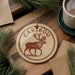 A circular ornament with a deer illustration and the word "CARIBOU" written on it, placed on a wooden surface next to a cup of coffee and pine branches.