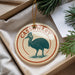 A circular ornament with a bird illustration and the word "CASSOWARY" written on it, placed in a box with pine branches.