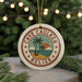 A round ornament with the text "CAVE CAUKER BELIZE" and a palm tree design, hanging from a string against a blurred background of a Christmas tree with lights.